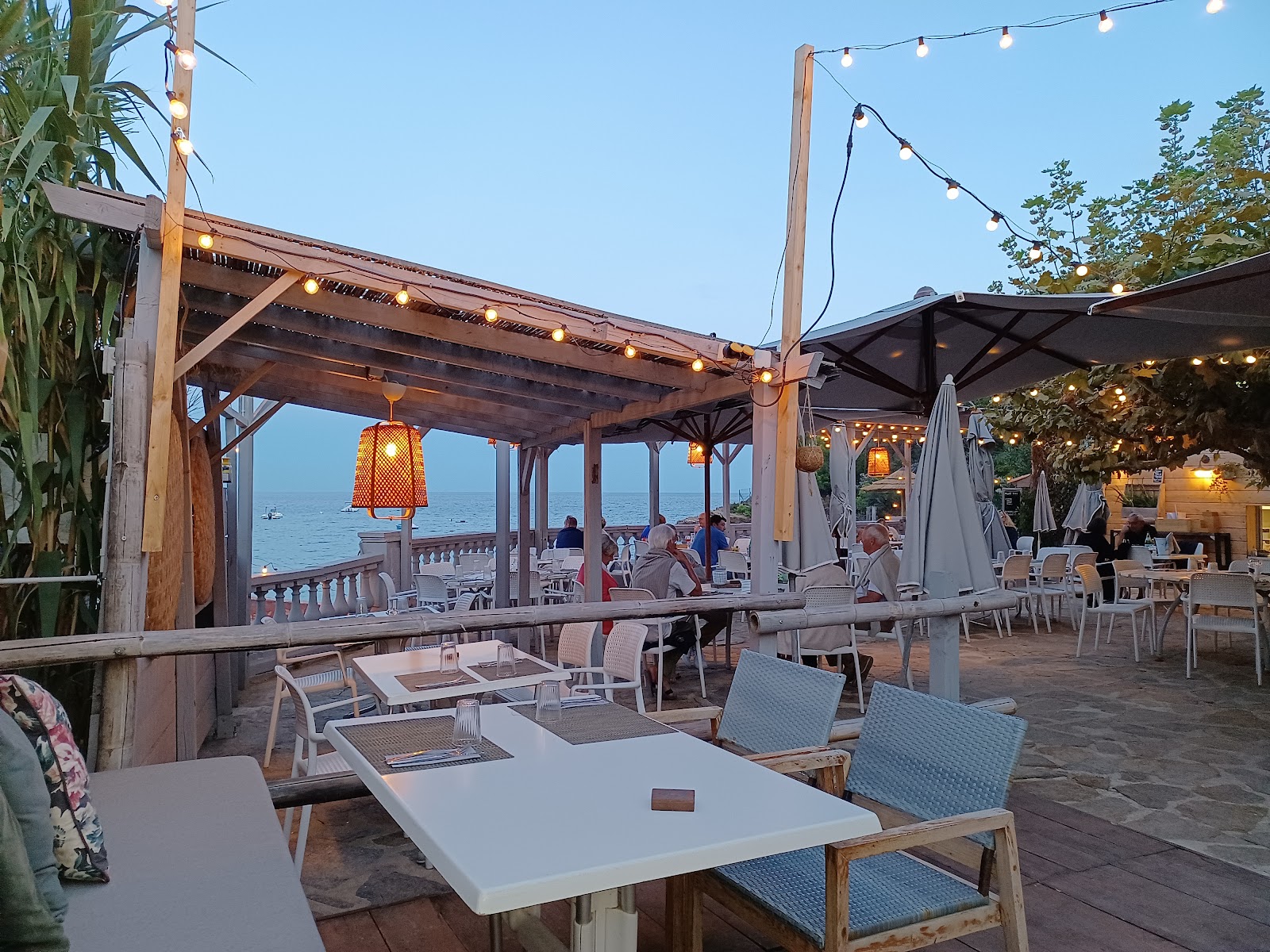 La terrasse d’Akwabô Beach au crépuscule : guirlandes lumineuses et lanternes orange sous la pergola, la mer calme au loin.