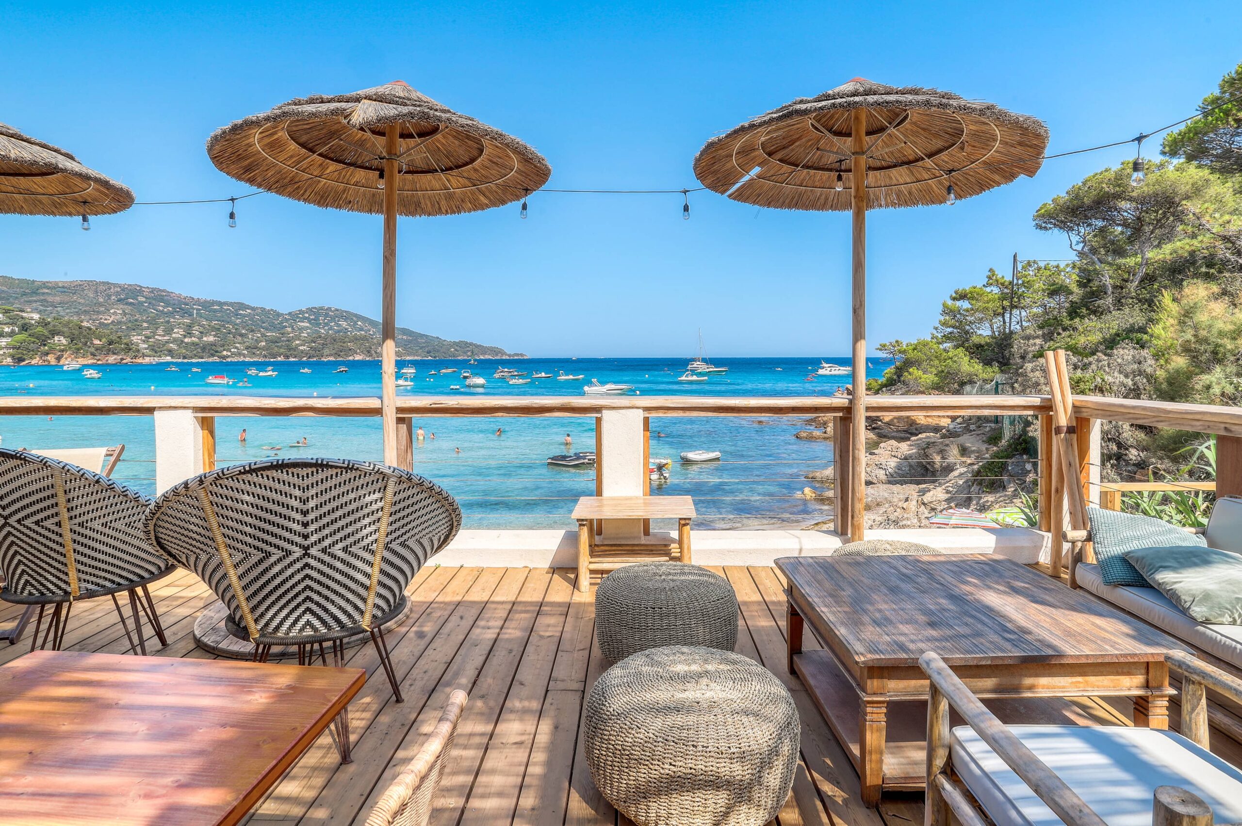 La terrasse en bois d’Akwabô Beach à Pramousquier : deux grands parasols de paille, fauteuils tressés, poufs, ouverte sur la mer turquoise et les bateaux.